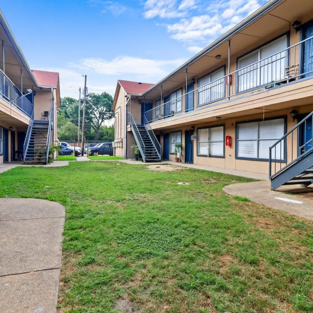 A view of the courtyard in an apartment complex, featuring two-story buildings with blue doors and multiple staircases, surrounded by a grassy area.