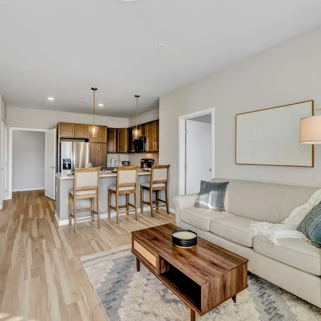 A stylish living room in a modern apartment, featuring a beige sofa with pillows, a wooden coffee table, and a woven storage unit. The space includes a kitchenette with dark wood cabinetry and barstools, and light wood flooring.