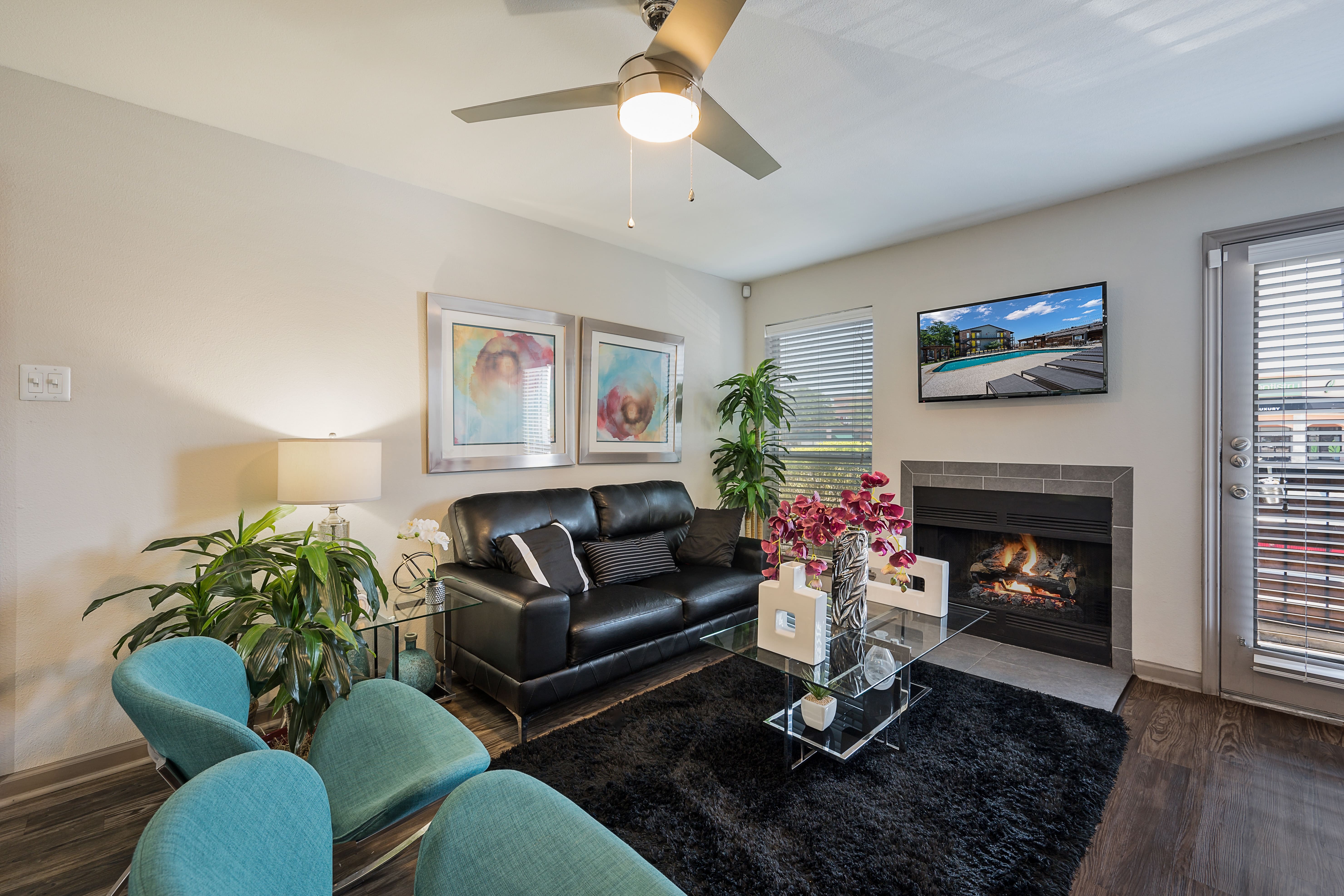 A modern living room featuring a black leather sofa, a glass coffee table, and a small fireplace. There are teal chairs and plants, with artwork on the walls and a TV mounted on the wall.