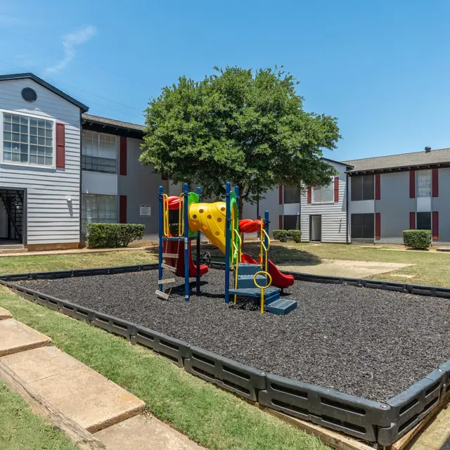 A playground area with a slide and climbing structure surrounded by rubber mulch, located in a spacious outdoor setting with grass and trees, near two apartment buildings.