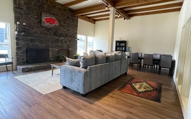 Spacious living room with a stone fireplace, wooden beams on the ceiling, a gray sectional sofa, and a dining area in the background.