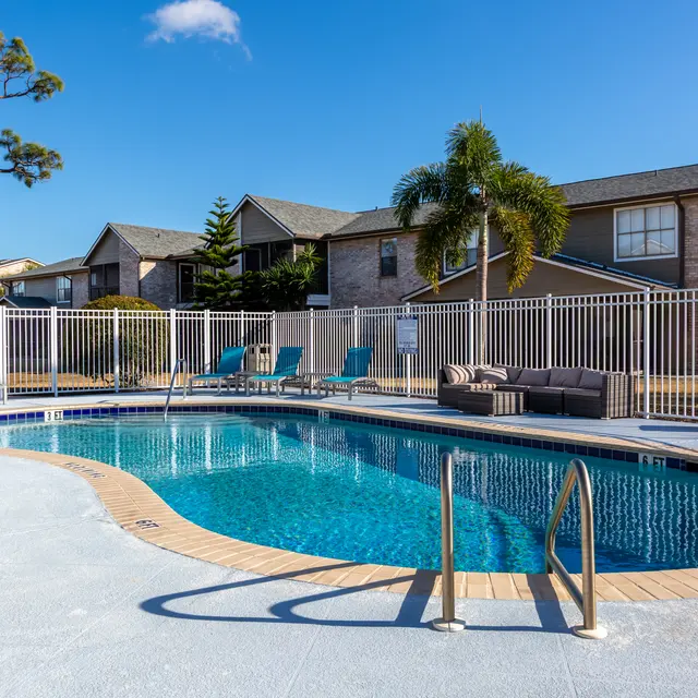 A bright, serene swimming pool area surrounded by lounge chairs and palm trees, with an apartment complex in the background.