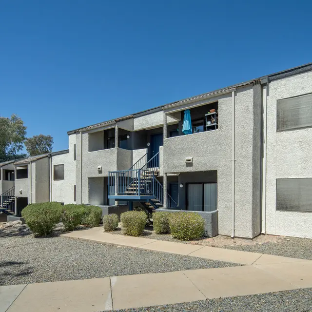 Two-story apartment building with balconies, surrounded by low shrubs and pathways, under a clear blue sky.