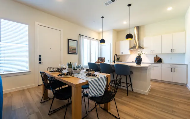 A modern kitchen and dining area featuring wooden flooring, a dining table with black chairs and decorative items, and a kitchen with white cabinetry and stainless steel appliances.
