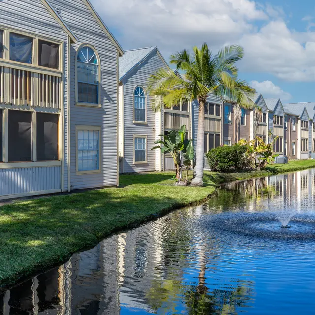 A serene view of a residential apartment complex beside a pond. The scene features multiple buildings with balconies, palm trees, and manicured grass along the water's edge, reflecting a blue sky with clouds.
