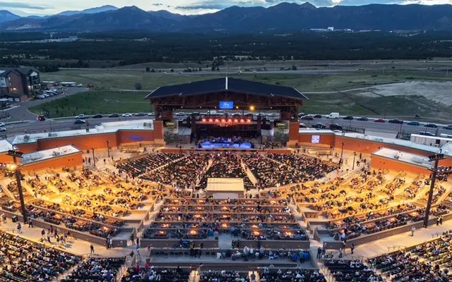 Aerial view of a large outdoor concert venue filled with an audience, illuminated by stage lights and surrounded by mountains.