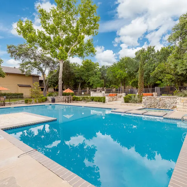 A view of a swimming pool surrounded by greenery and outdoor seating areas under a blue sky with scattered clouds.