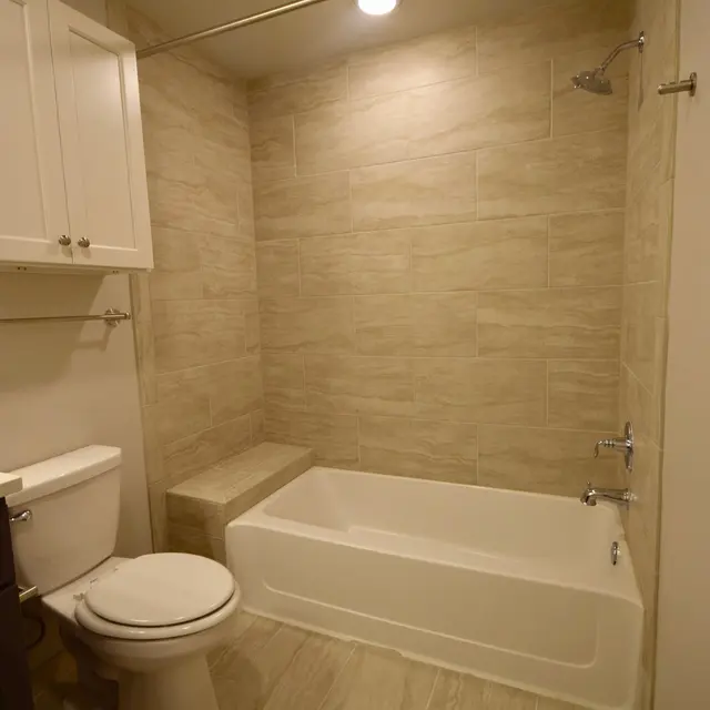 A modern bathroom featuring a white bathtub, shower area, and a toilet. The walls are tiled with light beige tiles, and there is a built-in bench in the shower area. The bathroom has a clean and minimalist design, with overhead lighting.