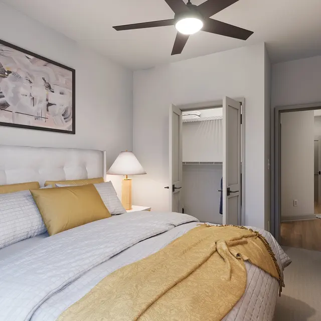A stylish modern bedroom featuring a large bed with gray and yellow bedding, a nightstand with a lamp, and a piece of wall art. There are two open doors leading to other rooms, and a ceiling fan above.