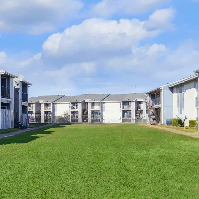 A wide view of a modern apartment complex featuring two-story buildings with balconies, surrounded by lush green grass and a clear blue sky with fluffy clouds.