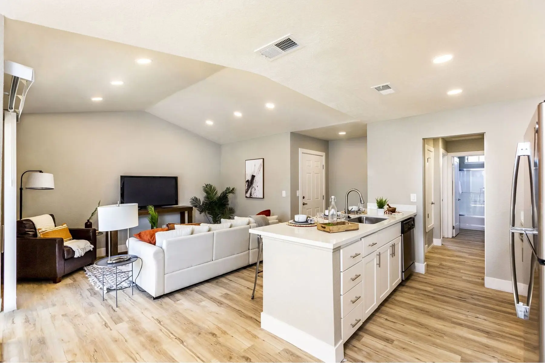 An open-concept modern living room and kitchen with light wood flooring. The living area features a brown leather sofa and a white sectional sofa, along with a coffee table and a green plant. The kitchen has a large island with seating and stainless steel appliances. The lighting is bright and airy, with recessed lights on the ceiling.
