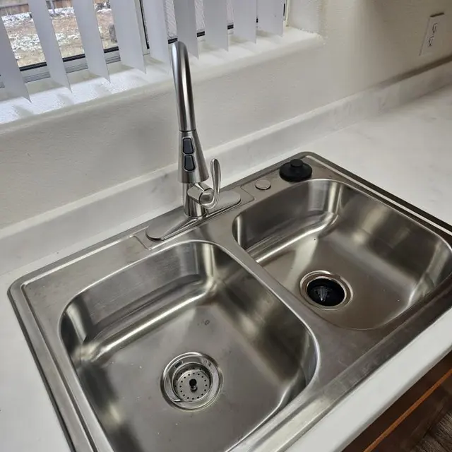 Modern Double Kitchen Sink A stainless steel double sink with a modern faucet in a kitchen. The sink is empty and features a drain in each basin, set against a light-colored countertop. Sunlight is streaming in through a window with vertical blinds.