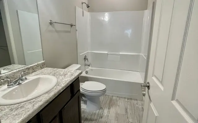 A sleek and modern bathroom featuring a white tub and shower combination, dark wood cabinetry, a single sink with a granite countertop, and light gray tiled flooring.