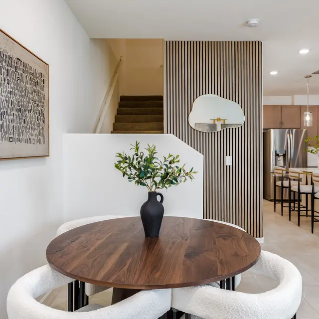 A cozy dining area featuring a round wooden table with white cushioned chairs, a vase of flowers on the table, and a modern decor style that includes abstract artwork on the wall and stylish light fixtures. Behind, there is a glimpse of a kitchen and staircase leading to another level.