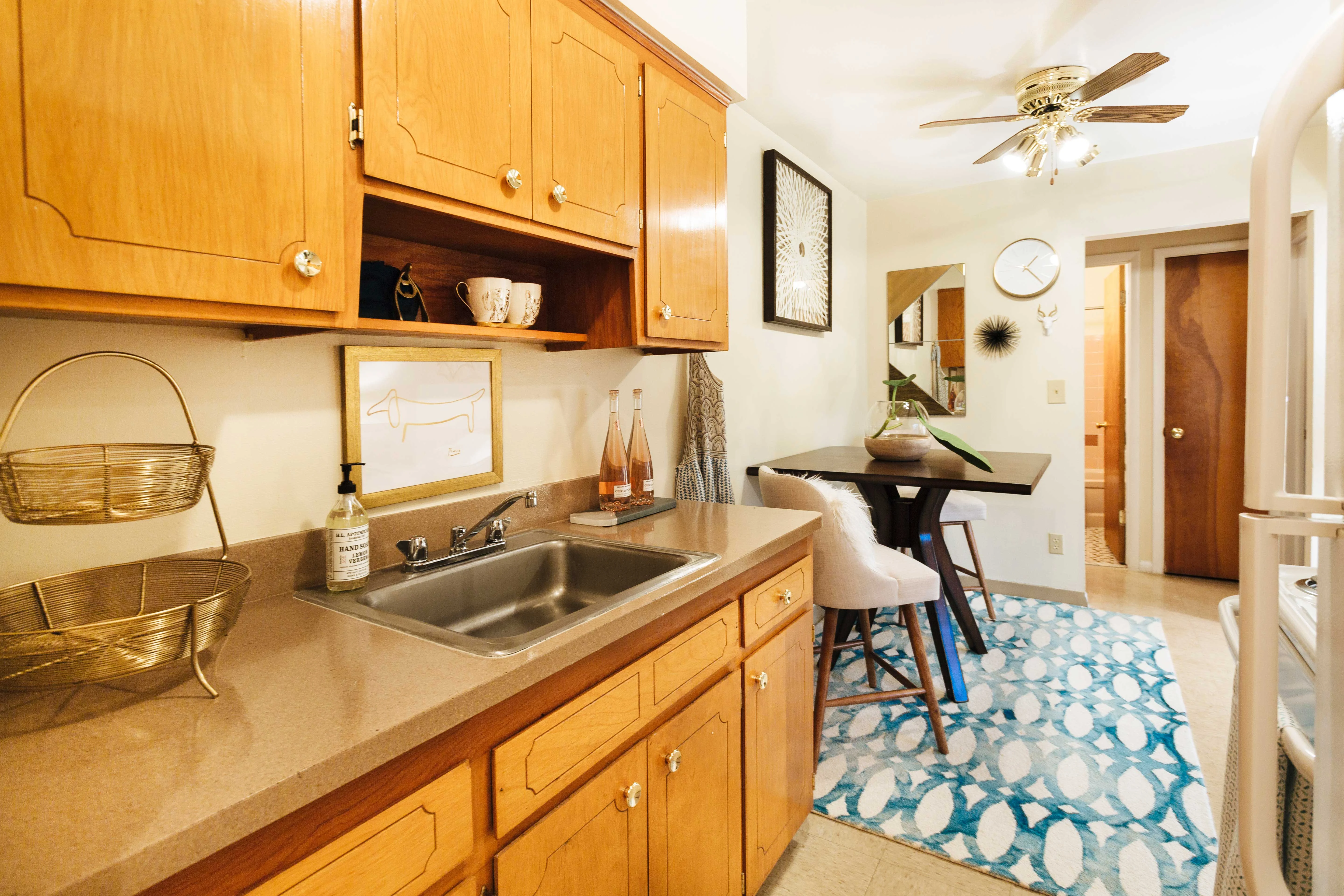 Cozy Kitchen Space A cozy kitchen space featuring wooden cabinets, a small sink area, and a dining nook. The counter is decorated with minimal kitchen accessories, and there is a patterned rug under a small table with white stools. A ceiling fan is visible above, and a wall clock hangs nearby.