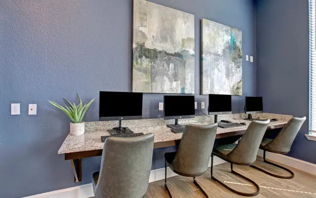 A modern office space featuring a row of computers with black monitors on a granite countertop, surrounded by stylish chairs. Abstract artwork adorns the walls, and there is a green plant for decoration.