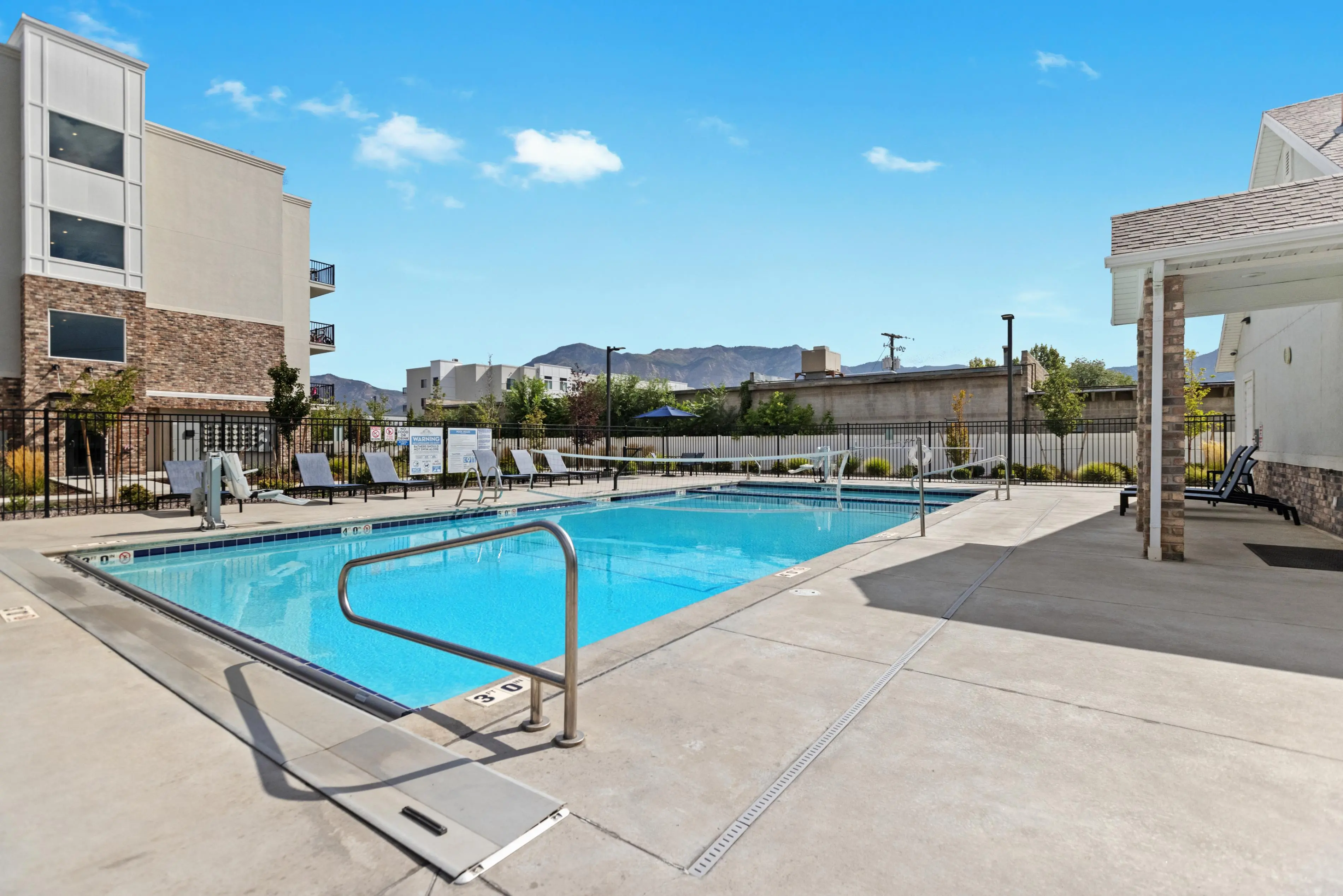 A clear blue swimming pool surrounded by lounge chairs and a modern building in the background under a sunny sky.