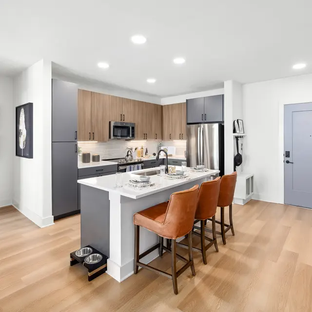 A modern kitchen with an open layout featuring a central island with four brown stools, stainless steel appliances, and light wood flooring. There are framed artworks on the wall and a door leading to the exterior.