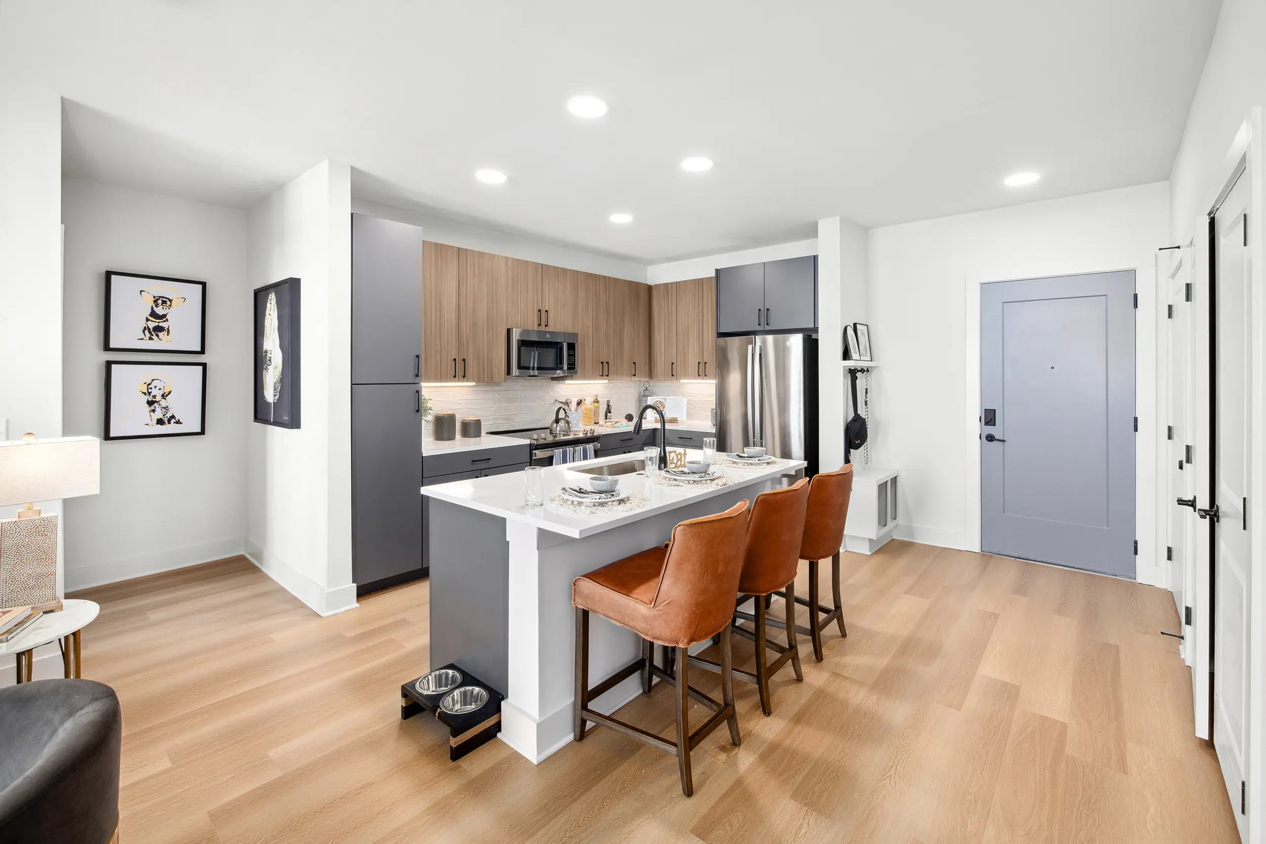 Stylish Modern Kitchen in Apartment A modern kitchen with an open layout featuring a central island with four brown stools, stainless steel appliances, and light wood flooring. There are framed artworks on the wall and a door leading to the exterior.
