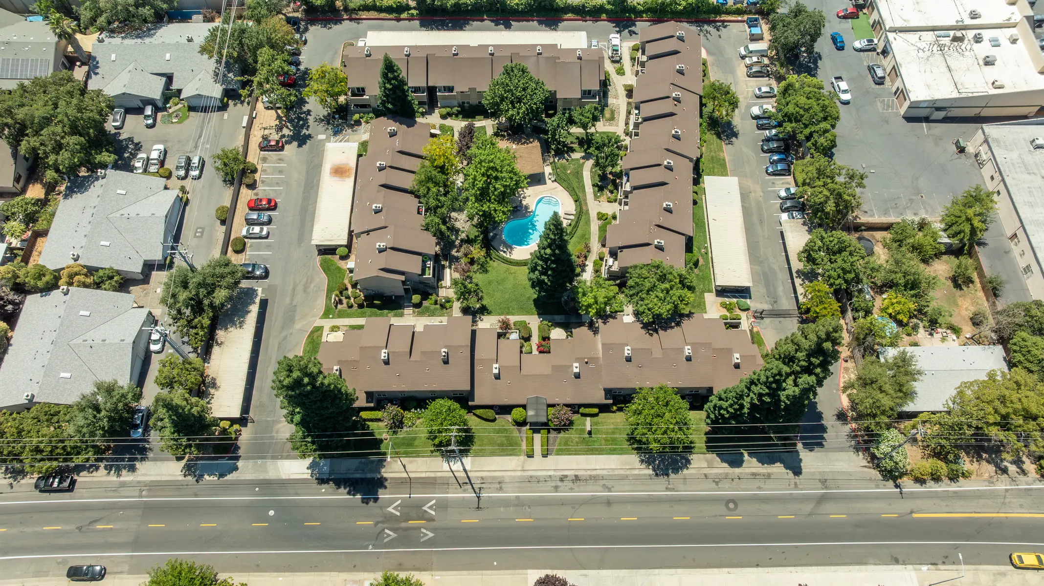 Aerial view of a residential apartment complex featuring brown-roofed buildings surrounding a central pool area and landscaped greenery.