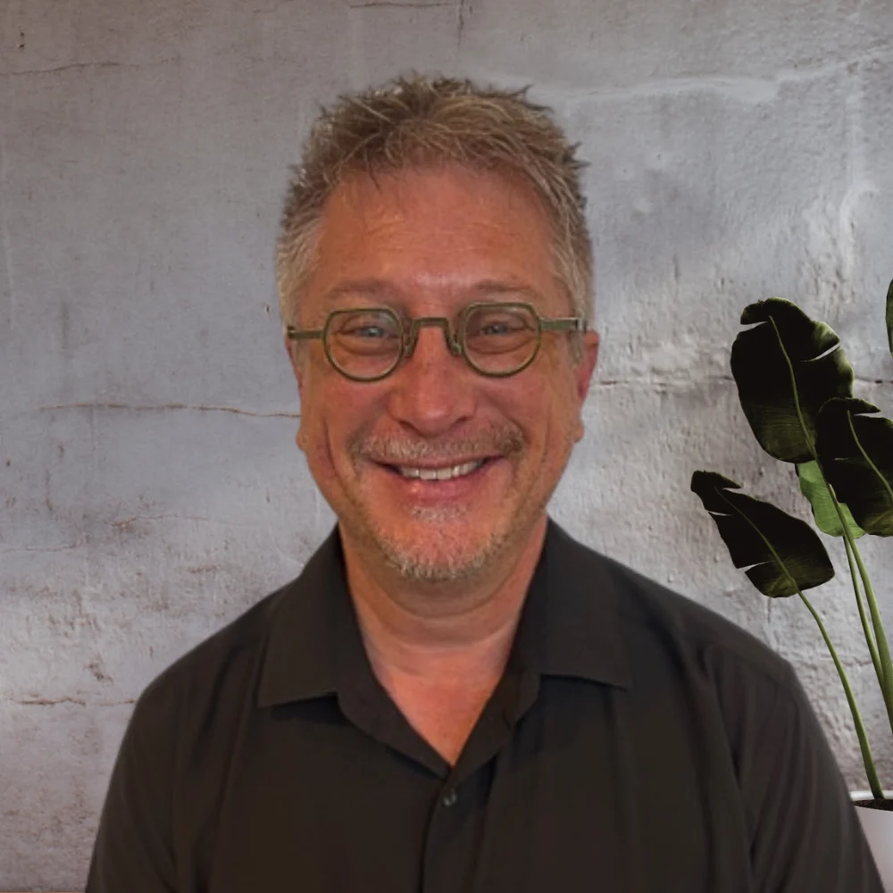 A man with short, light-colored hair, wearing glasses and a black shirt, smiles warmly at the camera. He is in front of a neutral background with a hint of greenery from a plant.