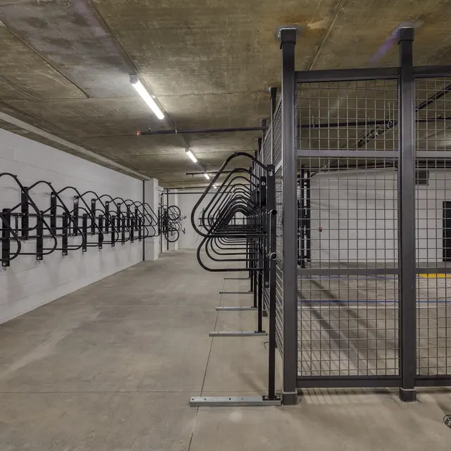A spacious underground bike parking area featuring wall-mounted bike racks on one side and a fenced-in section for bikes on the other side, with concrete flooring and exposed ceilings.