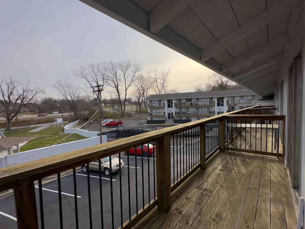 Balcony View Over Parking Area A wooden balcony with a railing overlooks a parking area and a tree-lined landscape. In the foreground, parked cars are visible, and in the background, a low-rise building can be seen along with bare trees.