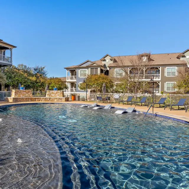 A sunny pool area at an apartment complex featuring a large swimming pool with lounge chairs and fountains, surrounded by trees and buildings.