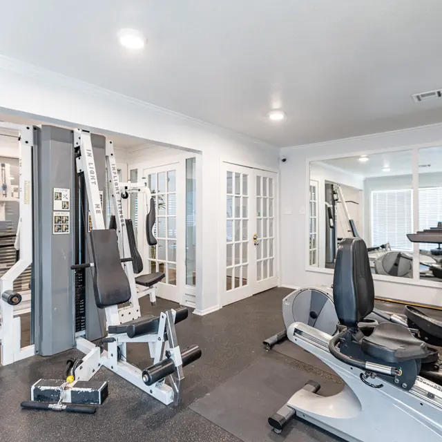 A spacious home gym featuring various workout equipment including a multi-station weight machine and cardio machines, with large mirrors and natural light coming from a window.
