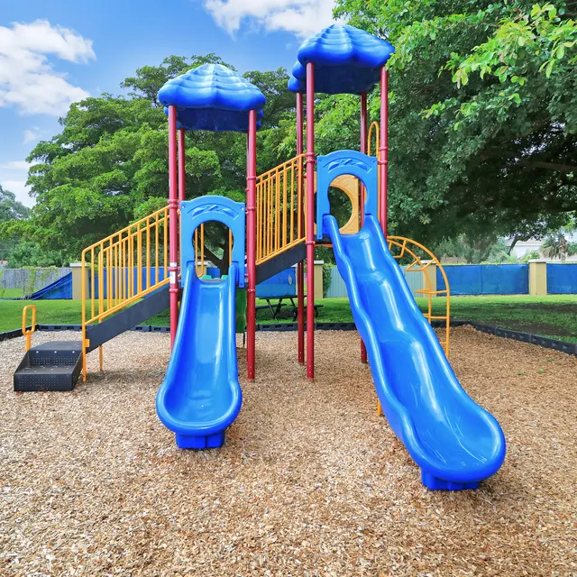 A colorful playground featuring two blue slides and a climbing structure, surrounded by grass and trees.
