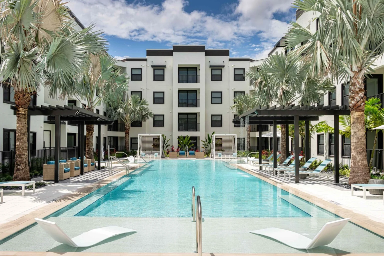 A modern apartment complex featuring a large pool surrounded by palm trees and lounge chairs.