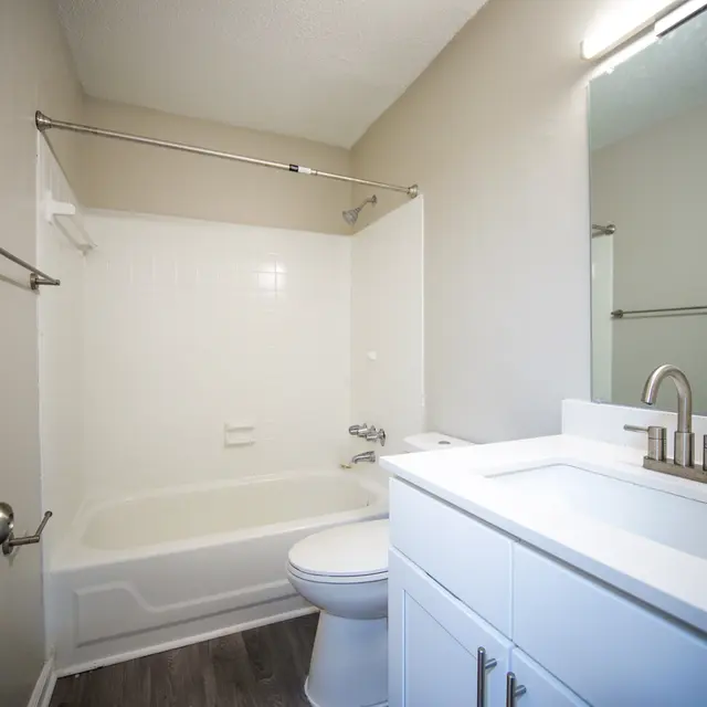 A clean, modern bathroom featuring a white bathtub, a white vanity with a sink, and a mirror. The walls are painted in a neutral color, and the floor is covered with brown vinyl planks.