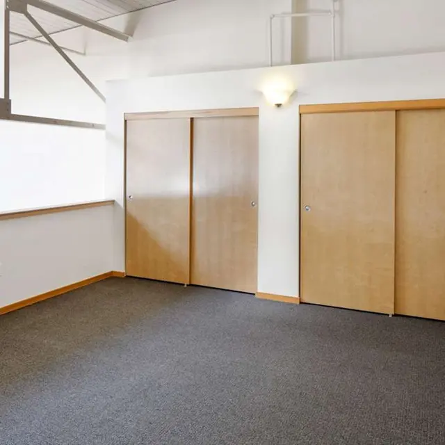An empty room featuring three wooden doors on one wall, with carpeted flooring and white walls, illuminated by soft lighting.