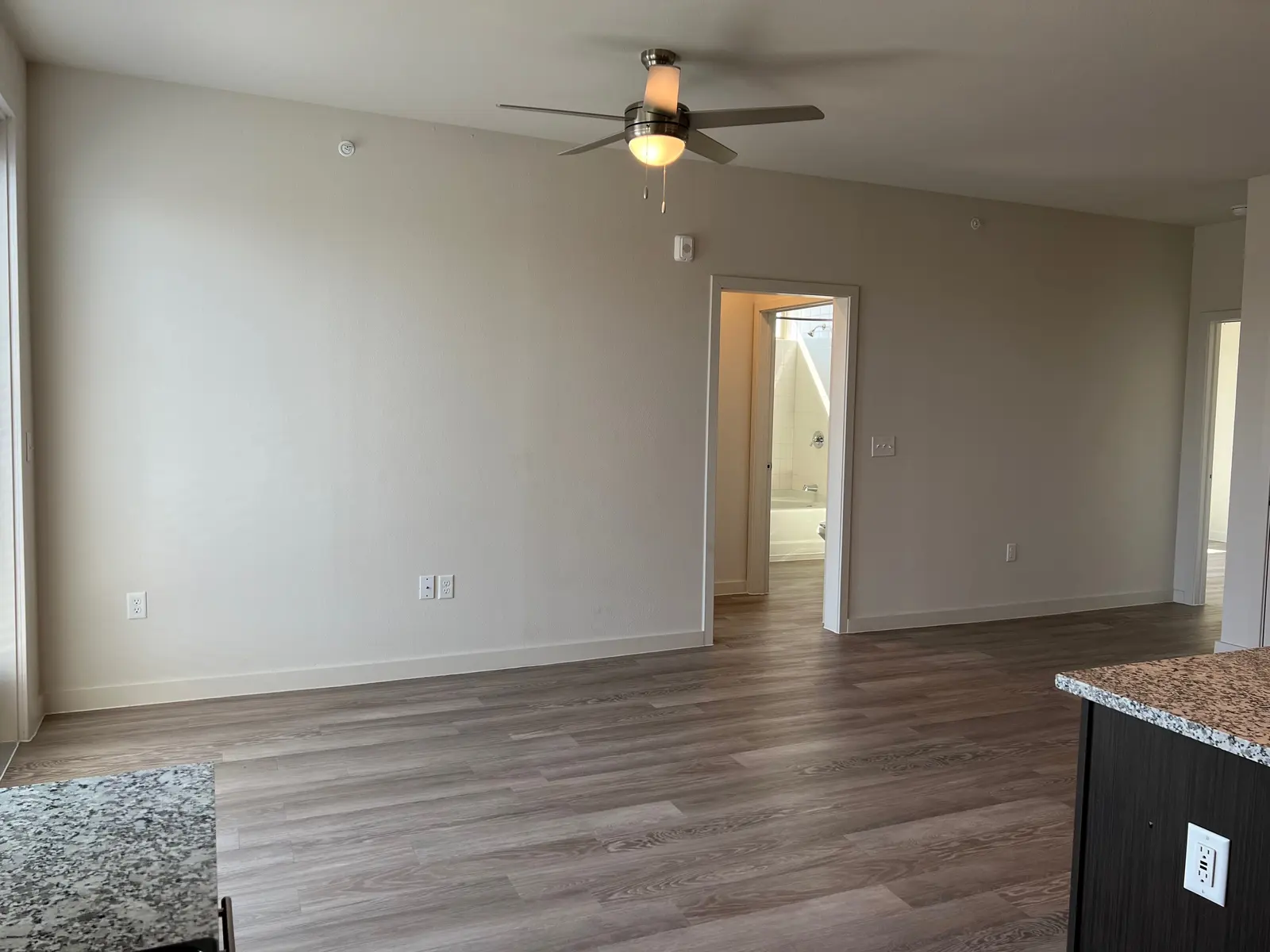 Spacious Living Area in Modern Apartment An empty living area featuring a ceiling fan, large windows, and a doorway leading to another room. The floor is wooden, and there is a granite countertop visible in the foreground.