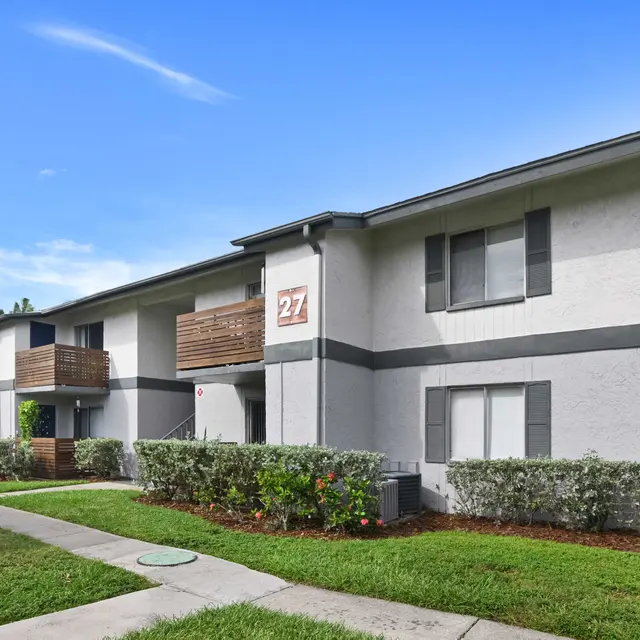 Exterior view of a two-story apartment building with the number 27 displayed, surrounded by greenery and a sidewalk.