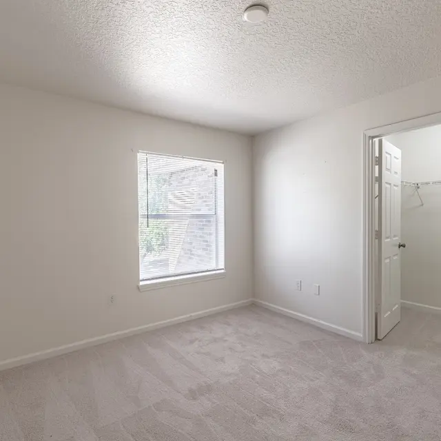 A spacious, empty bedroom with light-colored walls and plush carpeting. There is a window allowing natural light to enter, and a doorway leading to a small closet space. The room is well-lit and airy, giving it a calming feel.