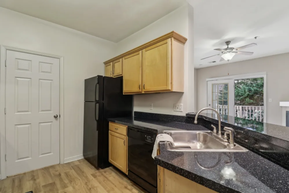 A modern kitchen with wooden cabinets, a black refrigerator, and a countertop. The kitchen features a sink and a view of an outdoor area through glass doors.