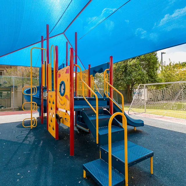 A colorful playground structure with slides and climbing elements, located under a blue shade cloth. A soccer goal is visible in the background on a grassy area.