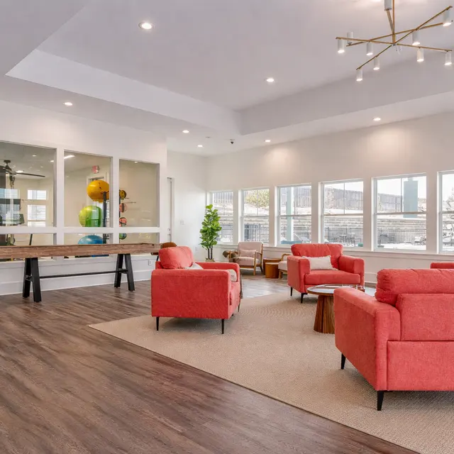 A spacious modern lounge area featuring red upholstered chairs, a wooden table, and large windows allowing natural light. There's a mix of seating arrangements and decorative elements like a potted plant and colorful balls visible in a separate room.