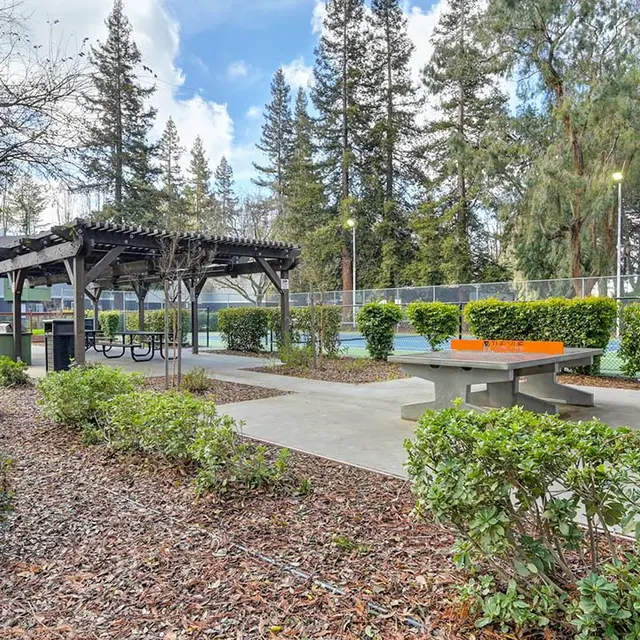 A park area featuring a sheltered seating structure and a ping pong table amidst greenery and trees.