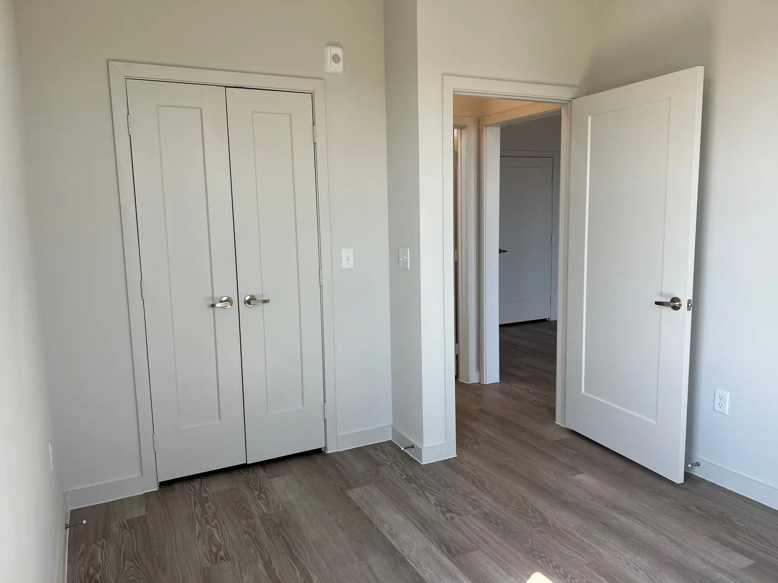 Empty Room Interior A corner in an empty room with light-colored walls, wooden flooring, and two sets of white double doors. One door is partially open, revealing another room. The lighting is bright and neutral.
