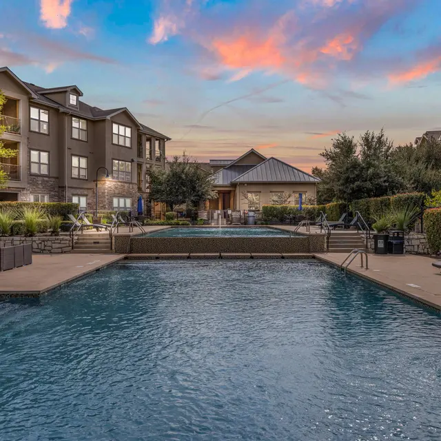 View of a swimming pool surrounded by lounge chairs and landscaped greenery, with residential buildings in the background under a colorful sunset sky.