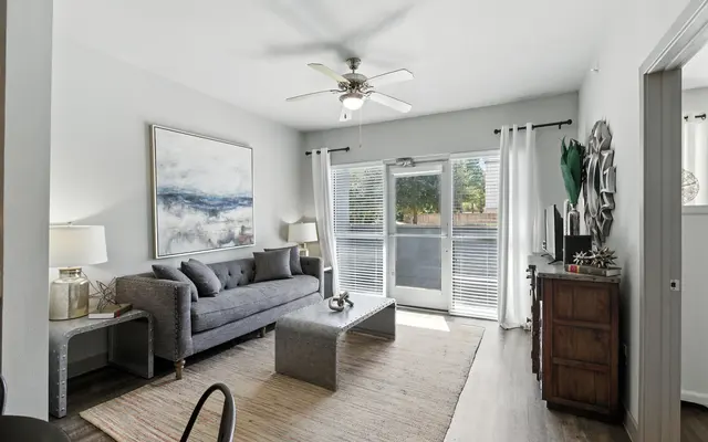 Modern living room with gray sofa, light walls, and large windows. Features a ceiling fan and sliding glass door to an outdoor view.