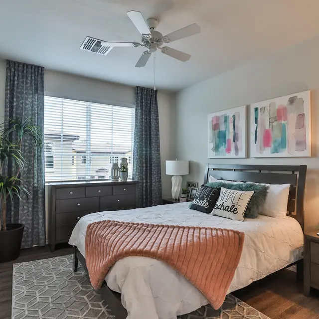 A modern bedroom featuring a queen-sized bed with a pink throw blanket, decorative pillows, and a stylish bed frame. The room has large windows with grey curtains, a plant in the corner, and modern artwork on the walls. Nightstands are placed on either side of the bed with lamps. The floor has a geometric patterned rug.