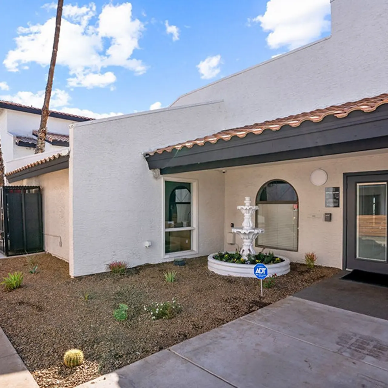 A modern apartment complex entryway featuring a small fountain surrounded by desert landscaping, palm trees, and bright blue skies.