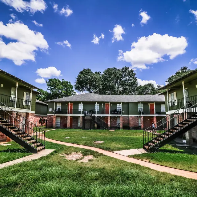 A view of a two-story apartment complex with green walls, visible staircases leading to the upper level, surrounded by grass and under a blue sky with fluffy clouds.
