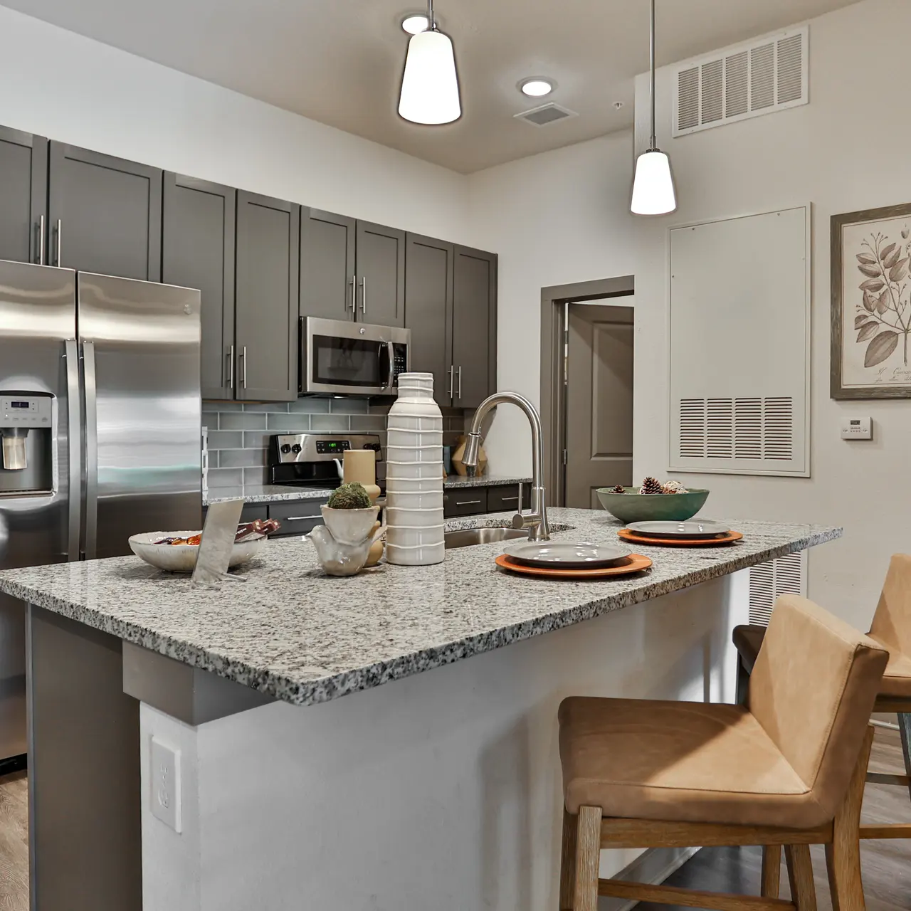 A modern kitchen featuring stainless steel appliances, a large granite countertop island with bar stools, and decorative vases on the counter. The cabinets are dark grey, and there are framed botanical artworks on the wall.