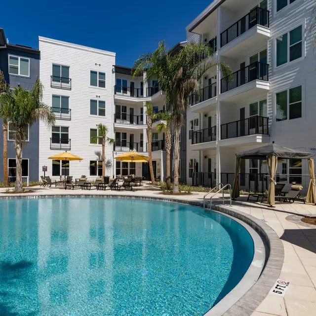 A sunny view of a modern apartment complex featuring a clear blue pool surrounded by lounge chairs and yellow umbrellas, with palm trees in the background.