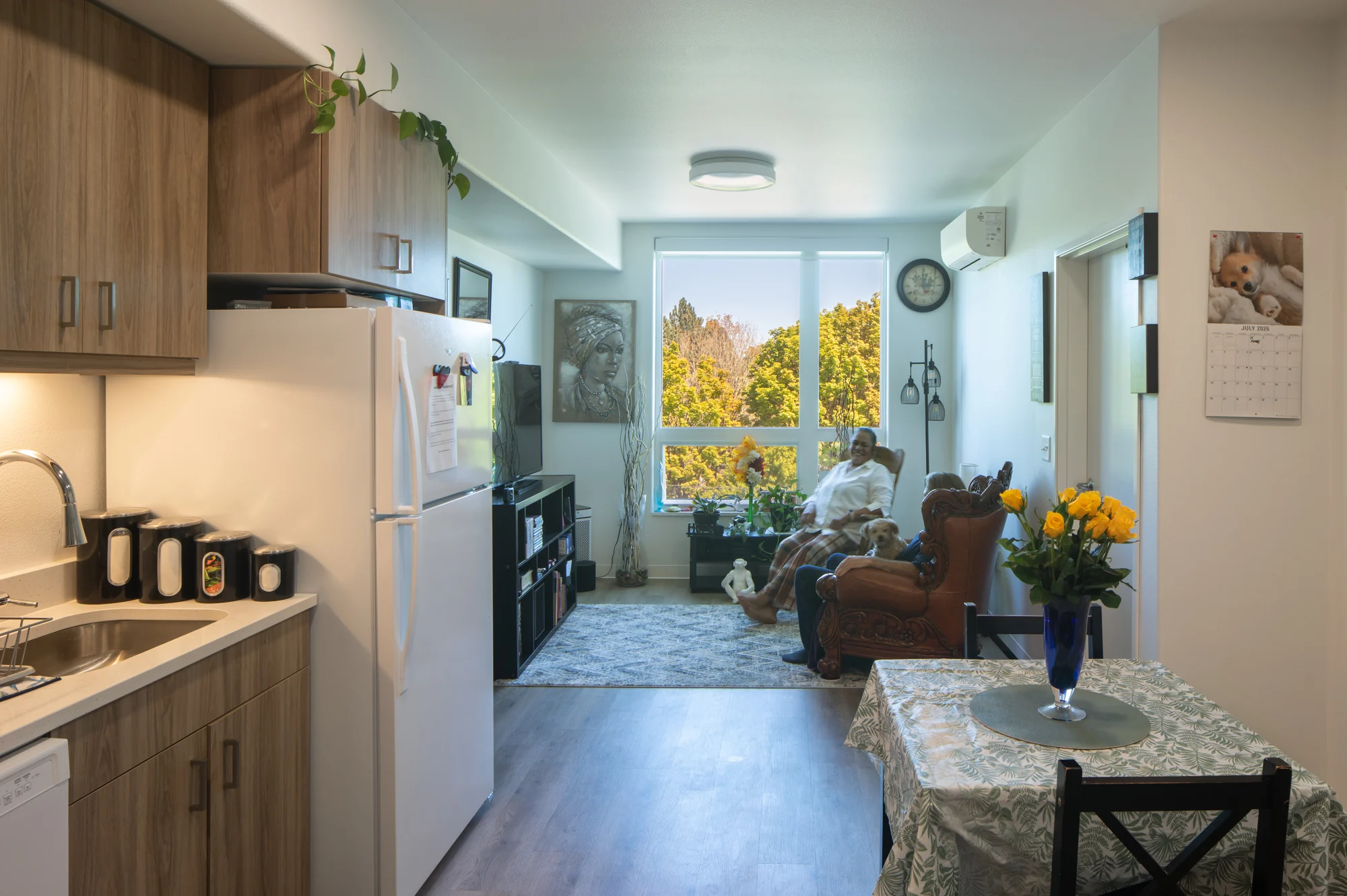 Cozy Apartment Interior A cozy apartment interior featuring a kitchen with wooden cabinets, a dining table with a floral centerpiece, and a living area with a man seated in an armchair beside a window.