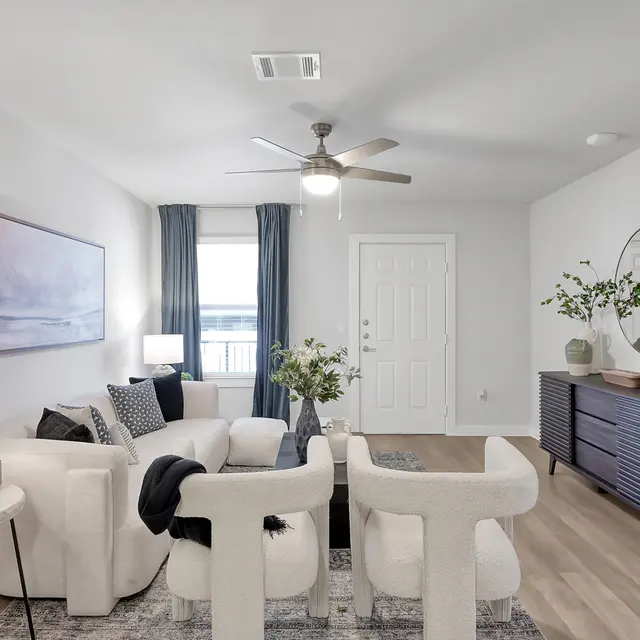 A modern living room featuring a white sofa with decorative pillows, a wooden coffee table, and a stylish dark wooden sideboard. The room is illuminated by natural light through a window, with blue curtains and tasteful decor including a mirror and plants.